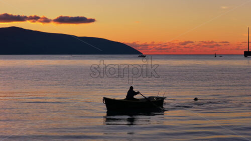Video - Aerial drone view of a lone fisherman rowing his small wooden boat during sunset in Vlora Bay, Albania, with soft orange skies, calm waters, and distant silhouettes of hills and clouds
