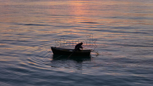 Video - Aerial drone view of a fisherman rowing a weathered wooden boat across the serene waters of Vlora Bay at dusk, creating gentle ripples that reflect the pastel evening light