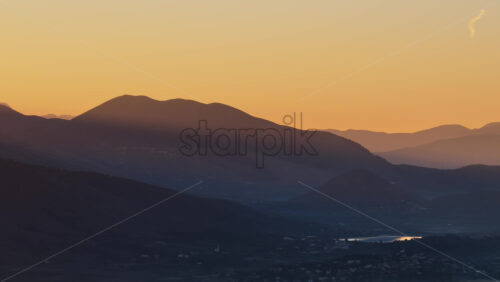 Video - Aerial drone view of a warm sunrise illuminating the layered mountain ridges in Albania, creating dramatic shadows and a soft golden gradient across the sky.
