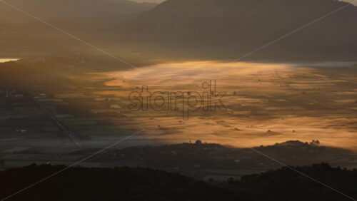 Video - Aerial drone view of morning fog drifting over the agricultural plains, glowing golden under early sunlight. Albania