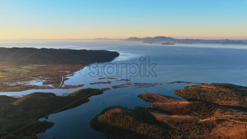 Video - Aerial drone view of Ksamil, Albania surroundings, revealing winding waterways, small islands, shallow wetlands, and the coastal landscape during warm sunrise light
