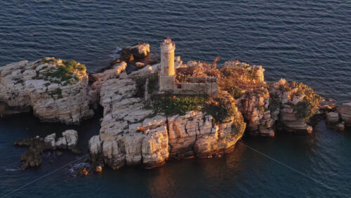 Video - Aerial drone view of an isolated rocky Peristeres Island featuring the old lighthouse ruins, illuminated by golden hour sunlight against the deep blue Adriatic Sea. Greece