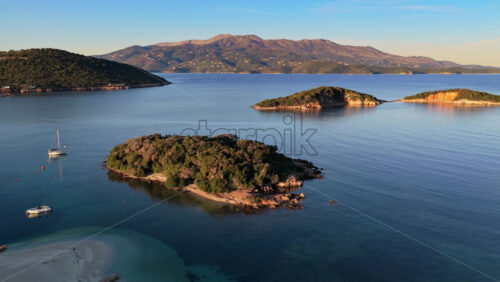 Video - Aerial drone view of the lush green Ksamil Islands surrounded by crystal clear waters, captured during early morning light with mountains in the distance