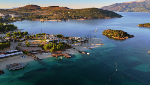 Video - Aerial drone view over Ksamil town, capturing its buildings, beaches, clear waters, and lush hillsides illuminated by soft sunlight