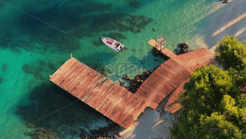 Video - Aerial drone view of a small white boat moored to a wooden pier above the transparent, emerald colored water of Ksamil