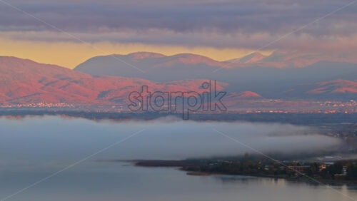 Video - Aerial drone view of the Ohrid, Macedonia region, showing fog lifting from the wetlands and soft pink light falling on the surrounding mountains and villages