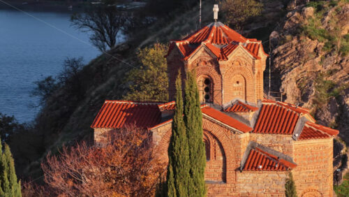 Video - Aerial drone view of the iconic St. John the Theologian Church perched above Lake Ohrid, framed by cypress trees and autumn colors. North Macedonia