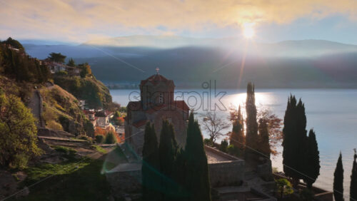 Video - Aerial drone view of a sunrise scene behind St. John at Kaneo, with rays breaking through the clouds and illuminating the church's silhouette. North Macedonia