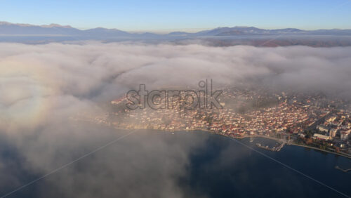Video - Aerial drone view of thick morning fog rolls over Ohrid's Old Town, partially revealing rooftops, the marina, and the surrounding mountains