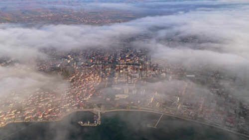 Video - Aerial drone view of Ohrid city partially obscured by drifting fog, with the marina and waterfront emerging from the mist while the mountains loom in the distance.