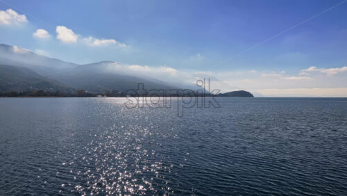 Video - Aerial drone view of the surface of Lake Ohrid, with the mountains gently covered in morning mist and a distant peninsula completing the peaceful landscape