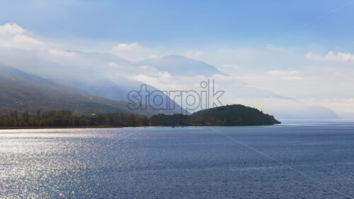 Video - Aerial drone view of the surface of Lake Ohrid, with the mountains gently covered in morning mist and a distant peninsula completing the peaceful landscape