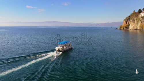 Video - Aerial drone view of a small passenger boat with a blue canopy glides across the deep blue waters of Lake Ohrid, leaving gentle ripples behind