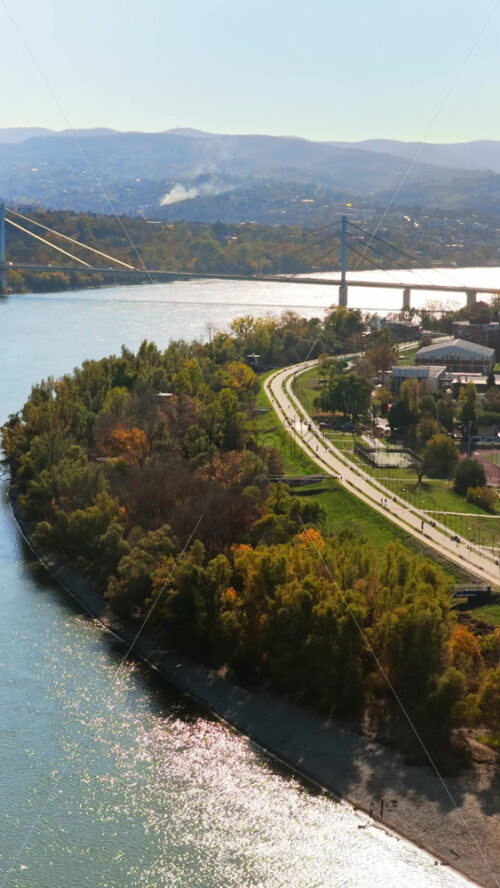 Video - Aerial drone view of the riverside promenade in Novi Sad, featuring the bike paths, sports courts, greenery, and the wide Danube River shimmering under the sun. Vertical