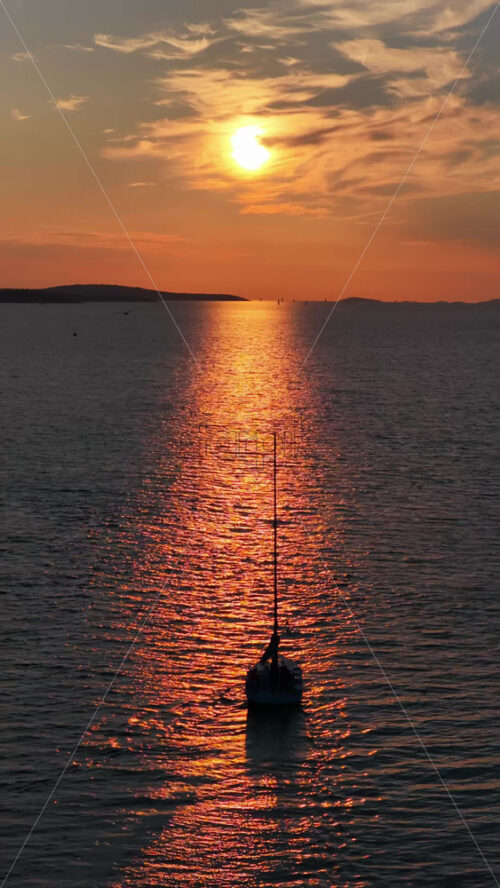 Video - Aerial drone view of a sailboat gliding on golden, sunlit water during sunset, with shimmering reflections surrounding the vessel. Vertical