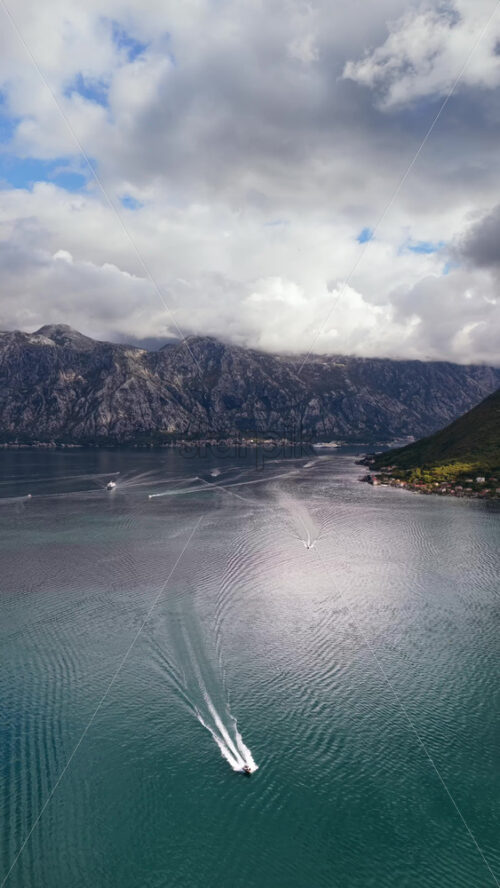 Video - Aerial drone view of speedboats leaving white trails on the calm water of the bay with mountains in the background. Vertical