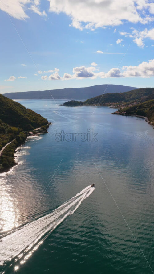 Video - Aerial drone view of a speedboat leaving white trails on the calm water of the bay with mountains in the background. Vertical