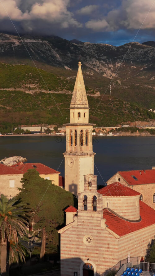 Video - Aerial drone view of Budva Old Town, showing its medieval stone buildings, red rooftops, fortress walls, and church tower along the coastline, with modern city buildings and mountains in the background. Vertical