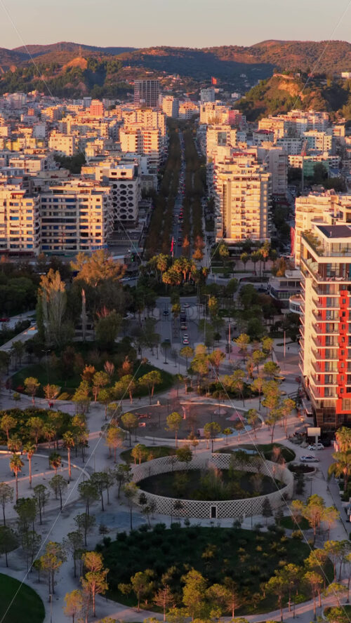 Video - Aerial drone view of the golden hour lighting highlighting Vlora's central urban park, featuring circular pathways, palm trees, playgrounds, and landscaped green spaces. Durres, Albania, Vertical