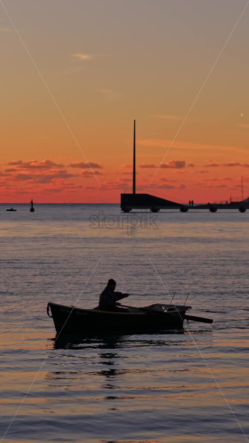 Video - Aerial drone view of a fisherman rowing a weathered wooden boat across the serene waters of Vlora Bay at dusk, creating gentle ripples that reflect the pastel evening light. Vertical