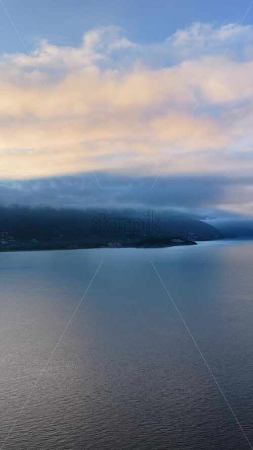 Video - Aerial drone view of Lake Ohrid, with low clouds hugging the mountains and soft pastel light reflecting across the water. Vertical
