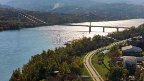 Video - Aerial drone view of the riverside promenade in Novi Sad, featuring the bike paths, sports courts, greenery, and the wide Danube River shimmering under the sun