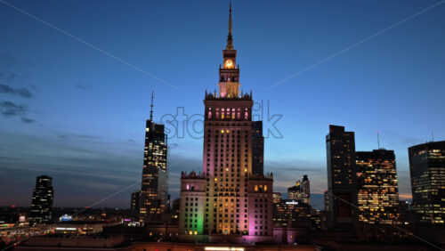 Video - Warsaw, Poland - October 30, 2025: Aerial drone view of the Palace of Culture of Warsaw and Science, glowing with colorful lights among the surrounding skyscrapers in the evening