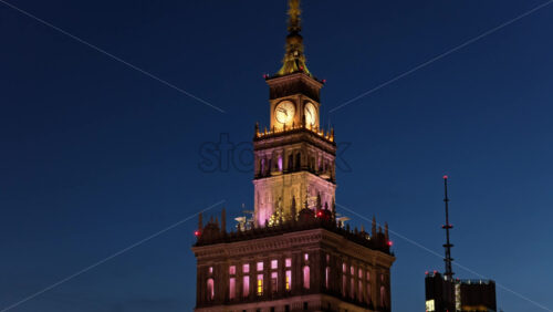 Video - Aerial drone view of the Palace of Culture of Warsaw and Science, glowing with colorful lights among the surrounding skyscrapers in the evening