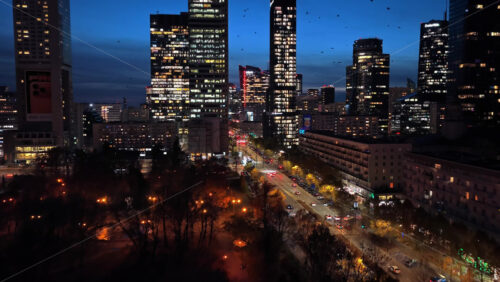 Video - Warsaw, Poland - October 30, 2025: Aerial drone view of busy nighttime streets full of trams, buses, and headlights, surrounded by high rise offices and shopping zones