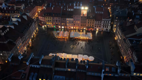 Video - Aerial drone view of Warsaw's Old Town Market Square, showing illuminated vendor stalls, people walking, and the geometrical layout of the historic square