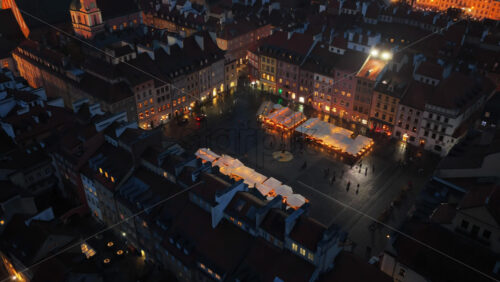 Video - Aerial drone view of Warsaw's Old Town Market Square, showing illuminated vendor stalls, people walking, and the geometrical layout of the historic square