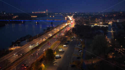 Video - Aerial drone view of Wybrzeze Gdanskie road along the Vistula River, showing heavy evening traffic, city lights, and the illuminated Swietokrzyski Bridge and National Stadium
