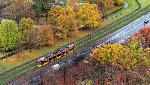 Video - Warsaw, Poland - October 30, 2025: Aerial drone view of a red and yellow tram traveling through green and orange autumn trees near Park Ujazdowski