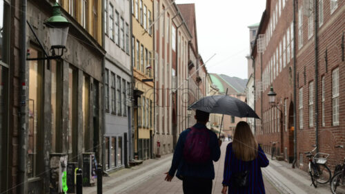 Video - Copenhagen, Denmark - August 3, 2025: Young couple walking together under an umbrella through old center city street on a rainy day