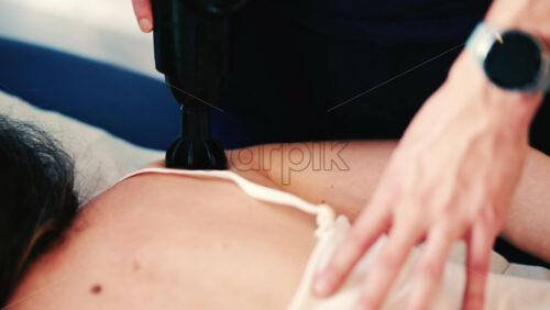Video - Therapist uses a percussion massage gun on a woman's shoulder while she lies on a treatment table