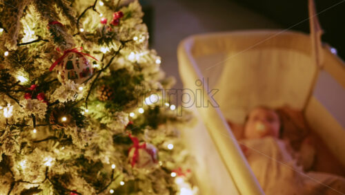 Video - Warm and intimate Christmas scene featuring a baby lying peacefully in a cradle beside a softly illuminated Christmas tree