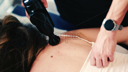 Video - Therapist uses a percussion massage gun on a woman's shoulder while she lies on a treatment table