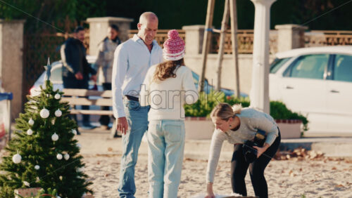 Video - Antibes, France - December 17, 2025: Group of people wearing winter clothing and Santa hats gathered around a decorated Christmas tree on a sandy beach taking family pictures