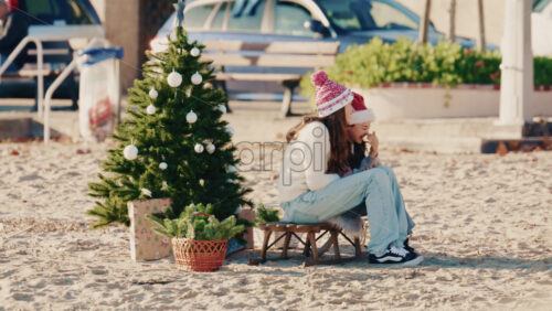 Video - Antibes, France - December 17, 2025: Young girls sitting on a small stool beside a decorated Christmas tree on a sandy beach taking pictures