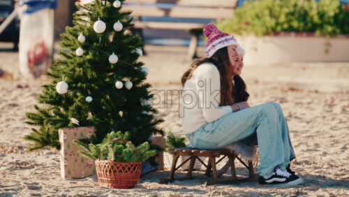 Video - Antibes, France - December 17, 2025: Young girls sitting on a small stool beside a decorated Christmas tree on a sandy beach taking pictures