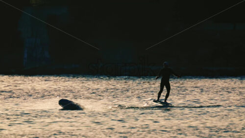 Video - Silhouette of a person riding an electric surfboard on calm sea water during golden hour