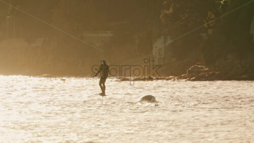 Video - Silhouette of a person riding an electric surfboard on calm sea water during golden hour