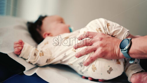 Video - Infant lying on a padded examination table while caregivers gently support the body during a routine check