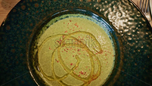 Video - Top view of a creamy green soup garnished with oil and spices, served in a ceramic bowl on a wooden table