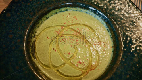 Video - Top view of a creamy green soup garnished with oil and spices, served in a ceramic bowl on a wooden table