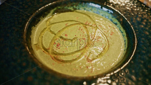 Video - Top view of a creamy green soup garnished with oil and spices, served in a ceramic bowl on a wooden table