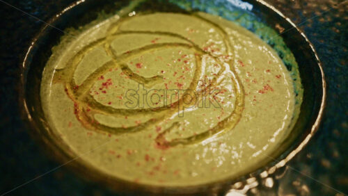 Video - Top view of a creamy green soup garnished with oil and spices, served in a ceramic bowl on a wooden table