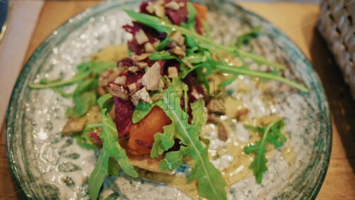 Video - Close up of a gourmet appetizer topped with fresh greens, nuts, and sauce, served on a ceramic plate