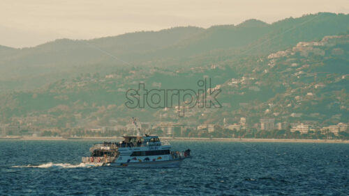 Video - Cannes, France - December 20, 2025: Passenger boat navigating the Mediterranean Sea with coastal hills and buildings in the background near Cannes, southern France