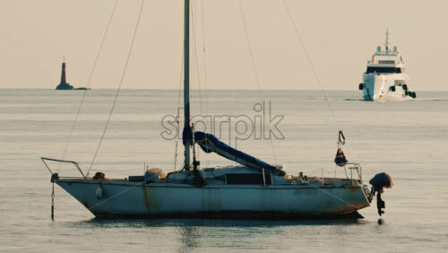 Video - A sailboat anchored on still water near the coastline, with a lighthouse and a large yacht visible in the background